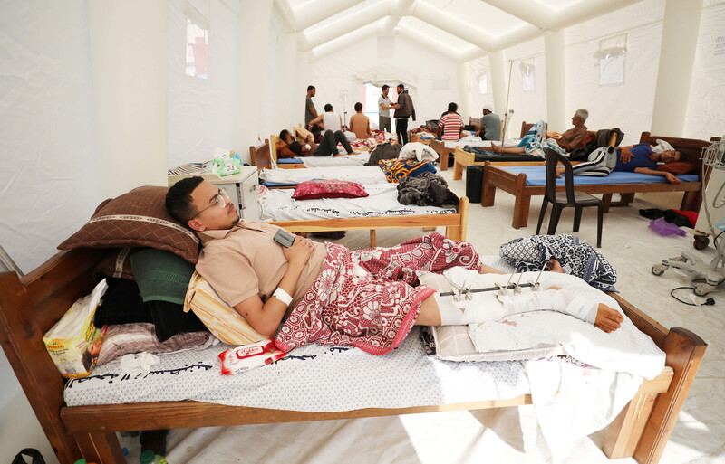 Young man reclines on bed in large tent lined up with additional beds occupied with patients