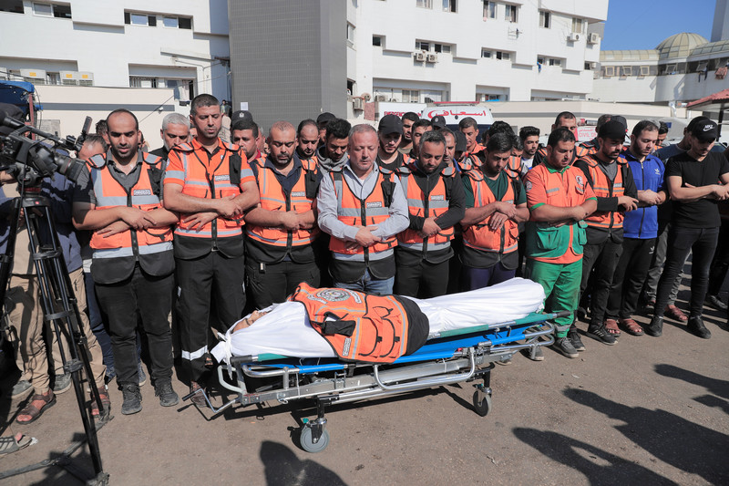 Men wearing orange safety vests stand with their arms crossed in front of their stomachs while standing in front of a shrouded body on a stretcher with a civil defense jacket placed on his chest
