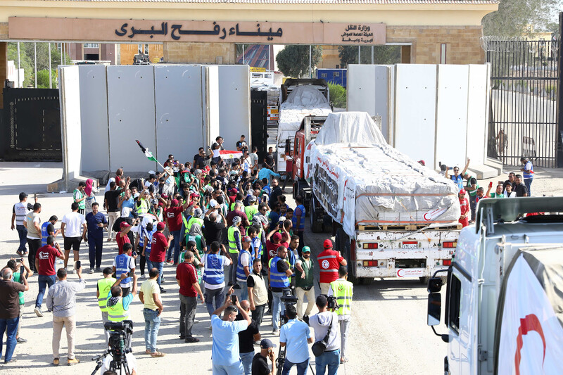 A crowd of people, many wearing safety vests, stand next to trucks carrying cargo at border crossing