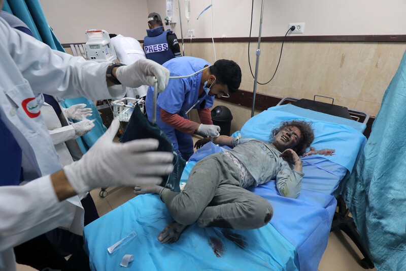 A man in scrubs leans over a girl covered in dust and blood lying on a hospital bed