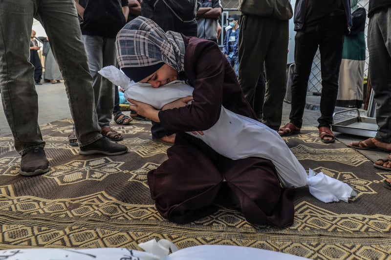 A young woman cries while holding her arms tight around a shrouded body and kneeling on the ground
