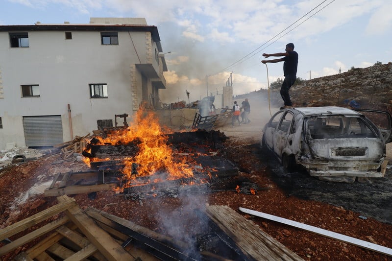 A fire blazes beside a burnt car amidst the destruction caused by a nearby building.