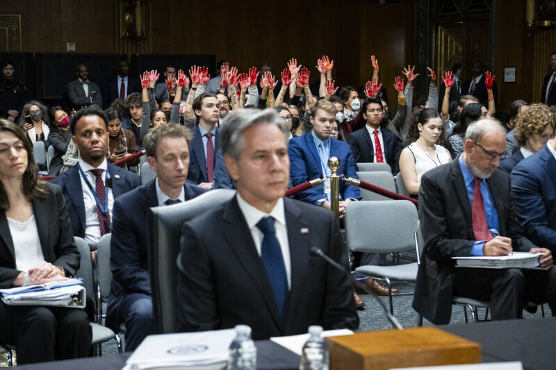 Man in a suit is seated while audience behind him has their hands raised and painted red 