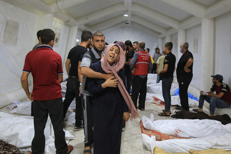 A man wraps his arm around a crying woman as they stand in a tent surrounded by shrouded bodies lying on the ground