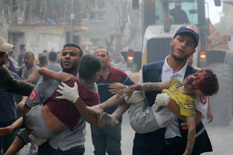 Two men carry children covered in dust and blood amid ruined buildings and chaos