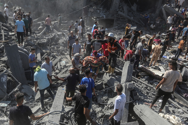 A man carries a body wrapped in a blanket as tens of people stand on rubble