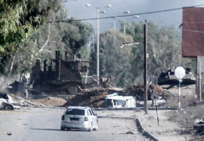 A sedan on a major road in front of Israeli military vehicles behind earth barriers