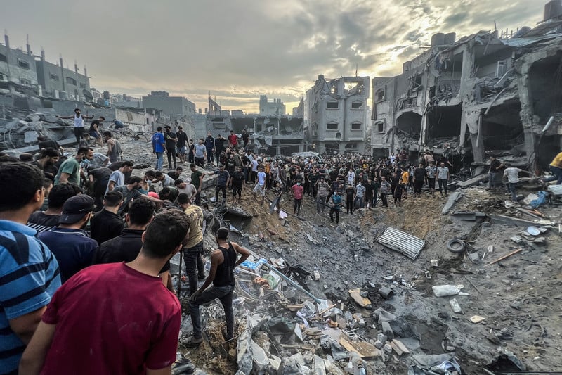 A crowd of men stand around a crater full of rubble surrounded by bombed-out residential buildings