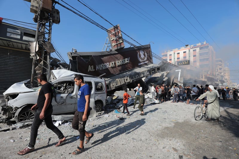People walk past the wreckage of a bakery and a car
