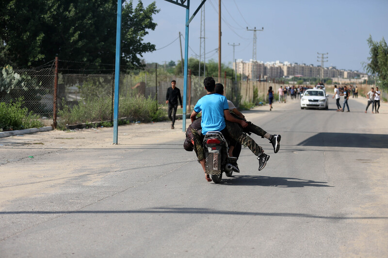 Two men are seen from the back riding a motorcycle with a man draped over the sides