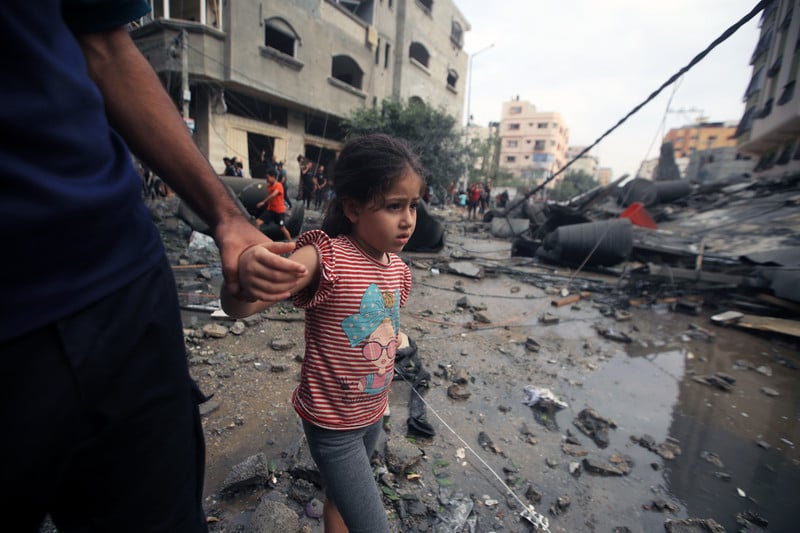 A girl holding an adult's hand walks along a rubble-strewn street