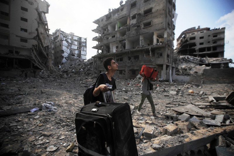 Young man looks up while holding a large roller suitcase as another person carries a suitcase in front of several bombed-out multi-story buildings 