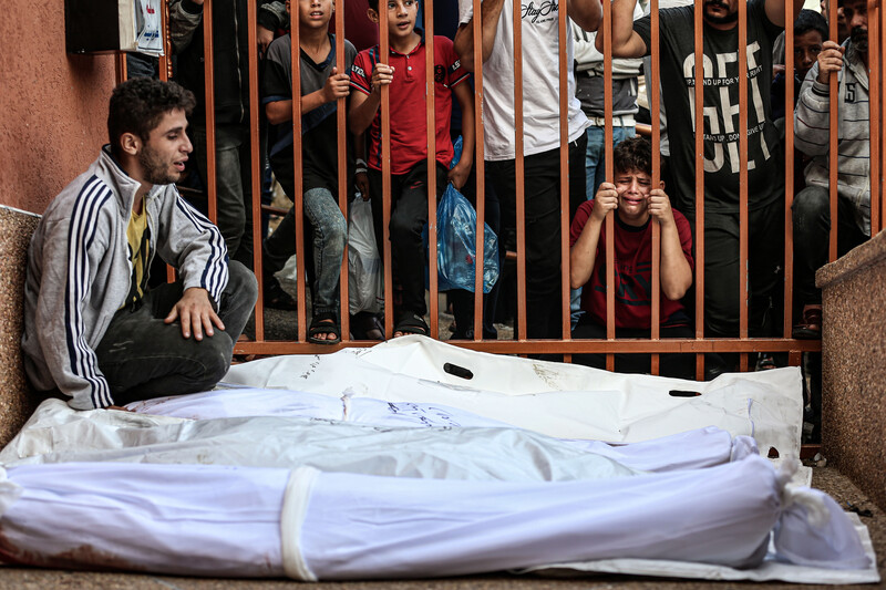 A man cries as he sits next to several shrouded bodies with mourning people standing on opposite side of metal fence behind him