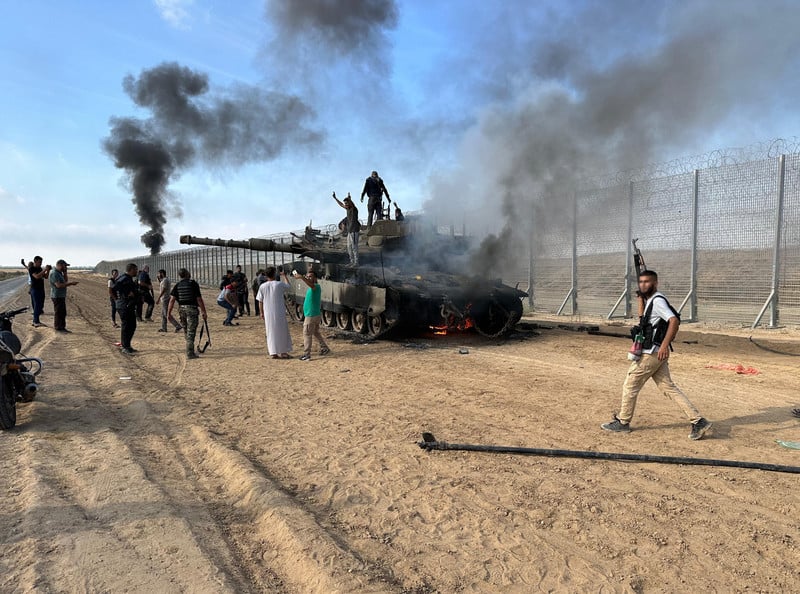 People climb on top of a burning Merkava tank as black smoke and flames rise from it