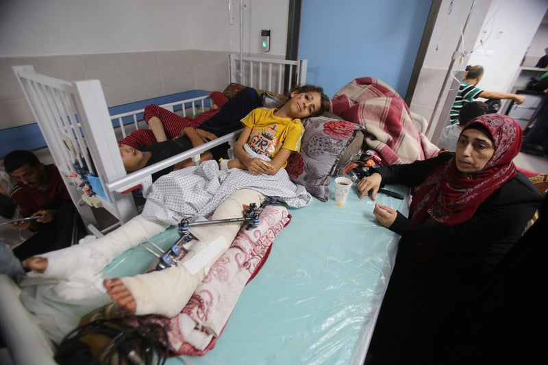 A girl with both of her legs bandaged and one in a splint rests on a bed with a woman sitting next to her