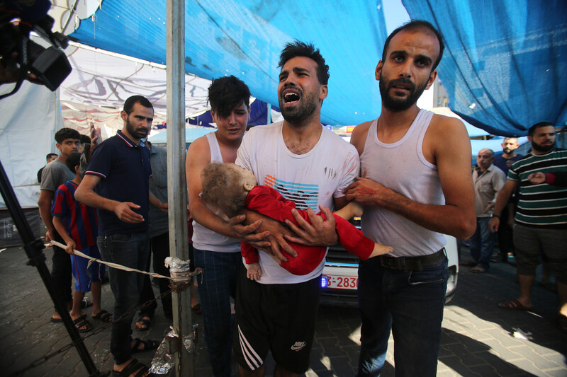 A young man with an agonized look on his face holds the body of a toddler while being embraced by two other men