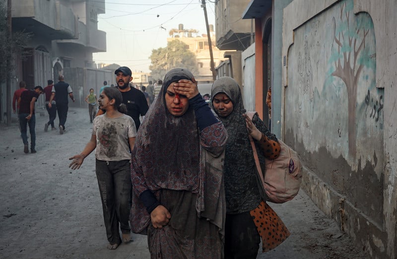 A woman with blood on her face holds her hand up to her forehead  while walking in a dusty alleyway with three other people