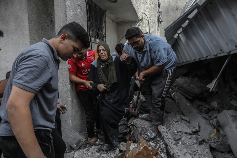 An older woman carrying a cane is supported by two young men as they walk through an alley filled with rubble