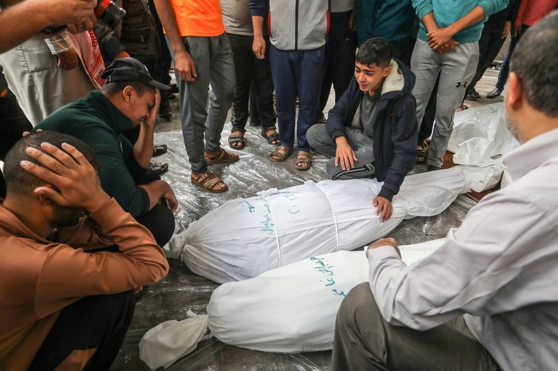 Palestinian men and a boy cry while sitting next to two shrouded bodies laid on a sheet of plastic