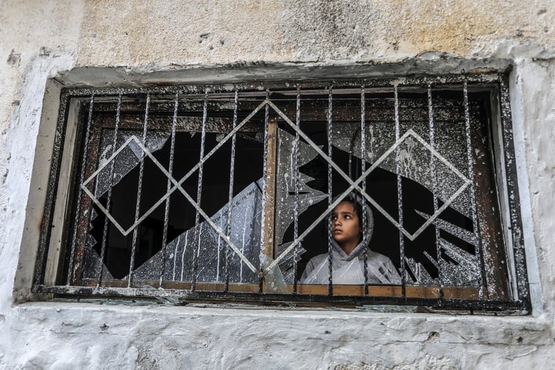 Girl looks out a broken window with blast marks