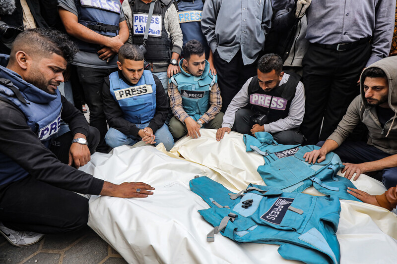 Several men wearing blue flak jackets marked as press stand and kneel next to two shrouded bodies with flak jackets placed on them