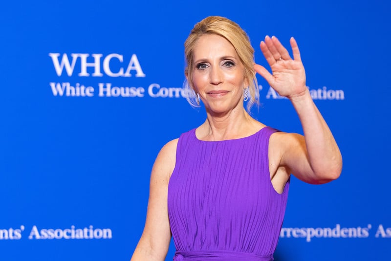 Dana Bash waves in front of a blue background with white lettering saying White House Correspondents' Association and WHCA