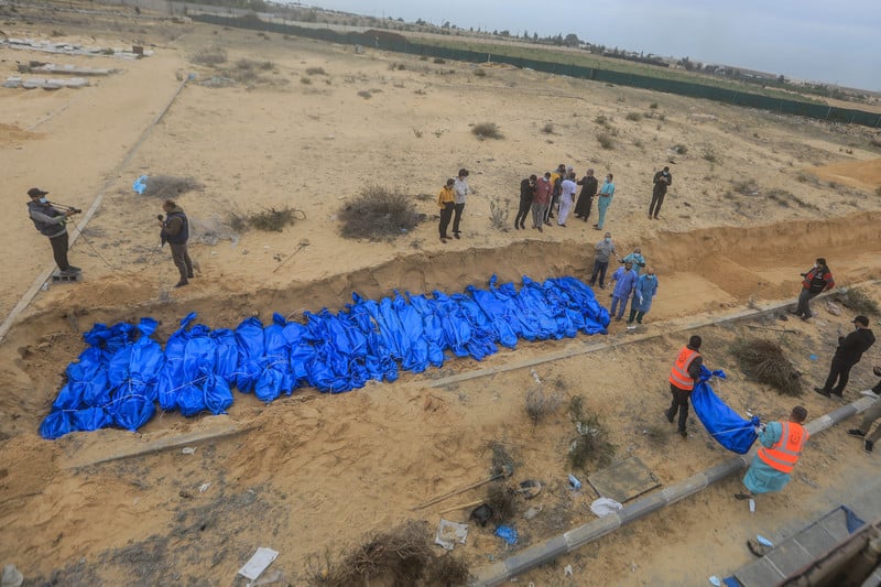 Aerial view of dozens of bodies wrapped in blue bags in a shallow trench with men standing near them