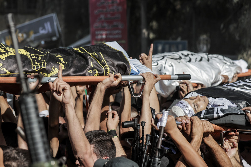 A view of hands carrying bodies wrapped in flags on stretchers while others hold rifles 