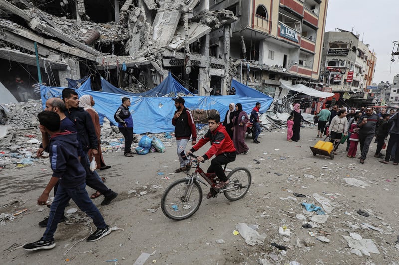 People walk and ride bicycles on a road with destroyed buildings from airstrikes