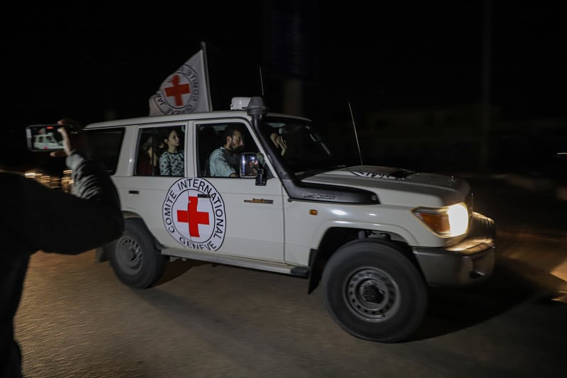 A Red Cross vehicle flying a white International Committee of the Red Cross flag with passengers drives on a dark road