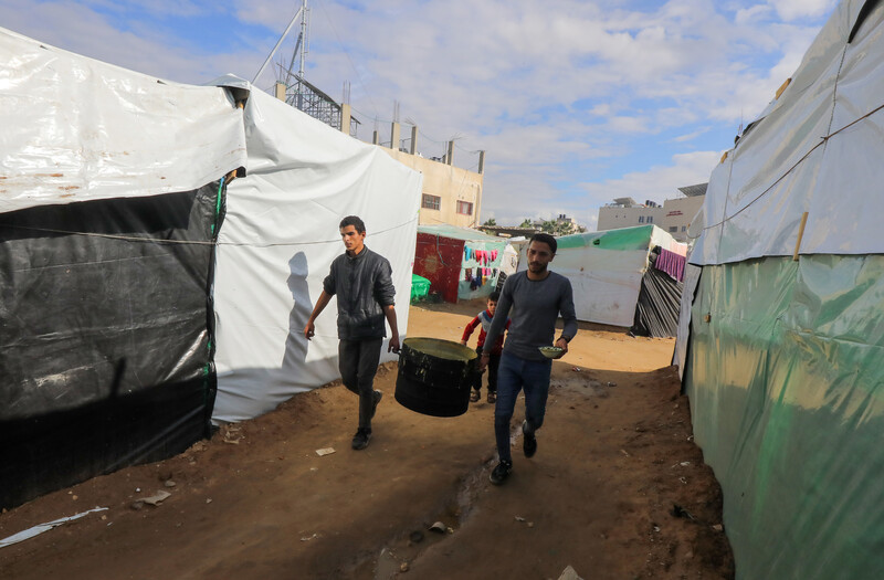 Two men carry a pot of soup cooked in a volunteer kitchen amid makeshift tents