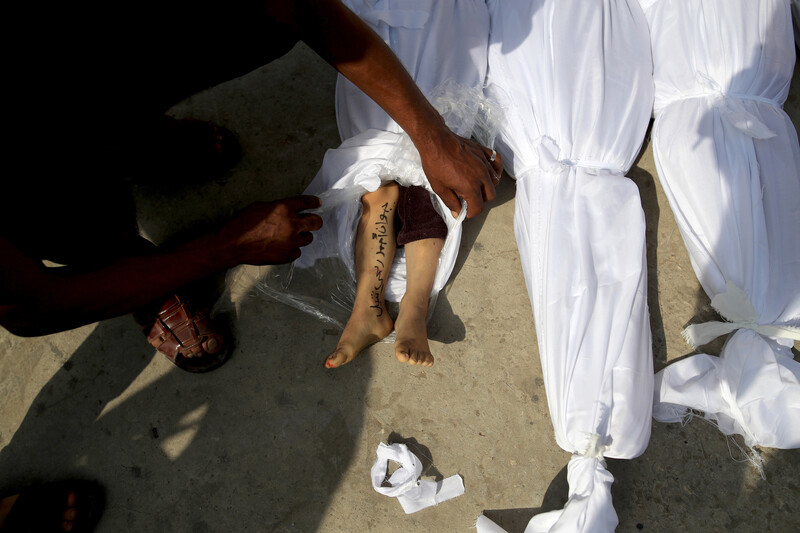 A person whose legs and arm are shown in the photo lifts a white sheet from the legs of a child's shrouded body