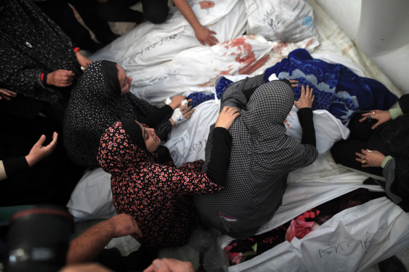 Women sit next to and lean over several bodies shrouded in white sheets