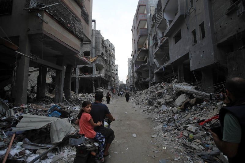 A girl on the back of a motorbike turns towards the camera while riding on a narrow road lined by bombed-out multistory buildings