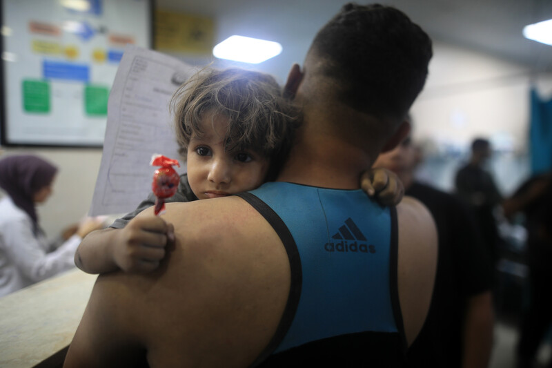 A man seen from the back carries a child who looks over his shoulders with wide eyes while clutching a lollipop