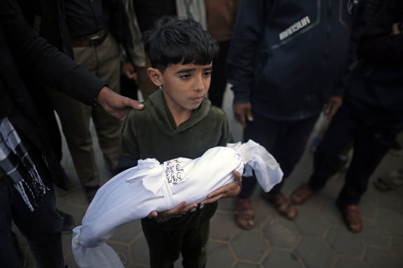 A young boy carries the body of a baby shrouded in a white cloth