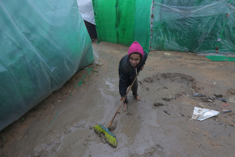 A small girl pushes a broom over mud beside makeshift tents