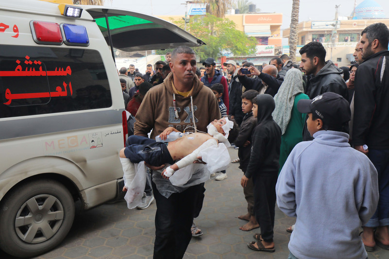 A man walks in front of an ambulance, carrying an injured child in his arms amidst a large crowd of people 
