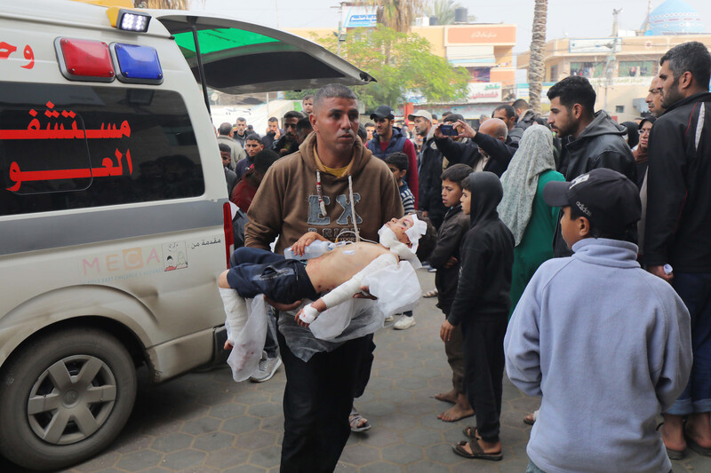 A man walks in front of an ambulance, carrying an injured child in his arms amidst a large crowd of people 