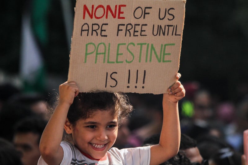 A child carries a sign that reads: None of us are free until Palestine is