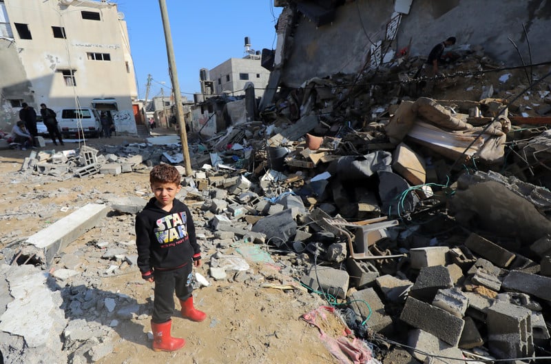 A young boy in a Star Wars sweatshirt and red rainboots stands next to a destroyed building
