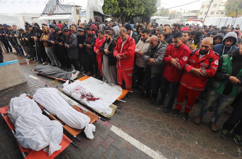 Mourners, some of them in red paramedic uniforms, pray over three bodies, wrapped in white shrouds.