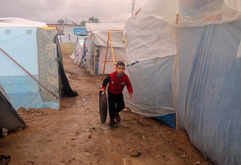 A boy carries a metal tray while walking on clay-like soil in between plastic-sheeted tents