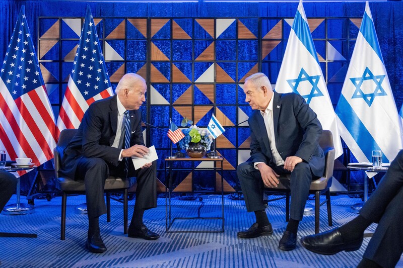 Joe Biden and Benjamin Netanyahu, both seated, lean toward each other with American and Israeli flags behind them