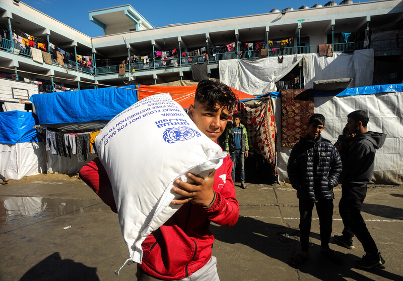 A Palestinian youth carries a bag of UNRWA flour with two youths and an UNRWA school in background