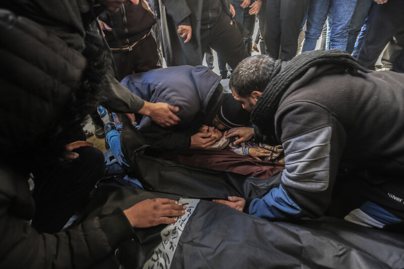 A kneeling man cradles and puts his head next to the face of a small child in a body bag as other men kneel and stand around him