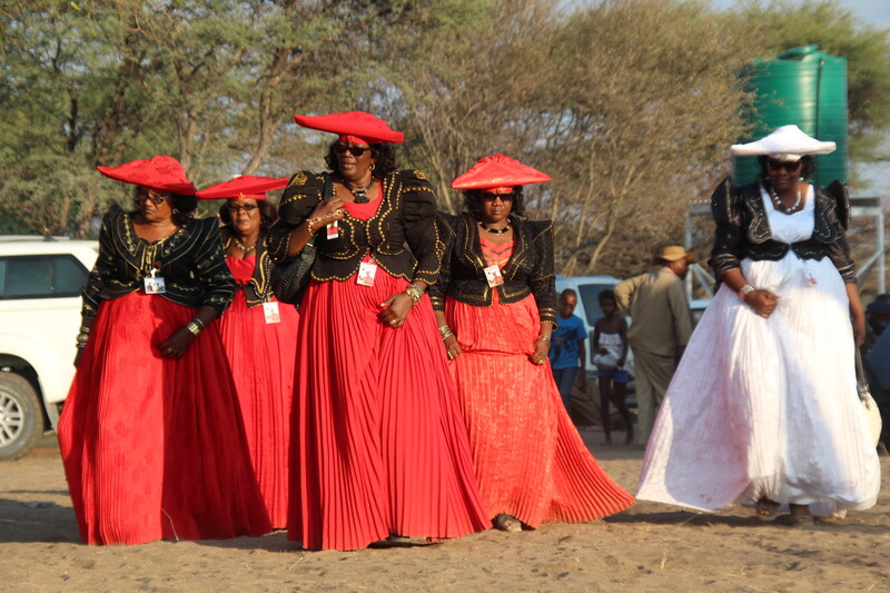 A group of women in bright flowing dresses and hats walk amid trees 