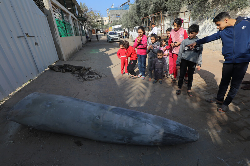 A group of young children in a street point at a large bomb lying on the ground