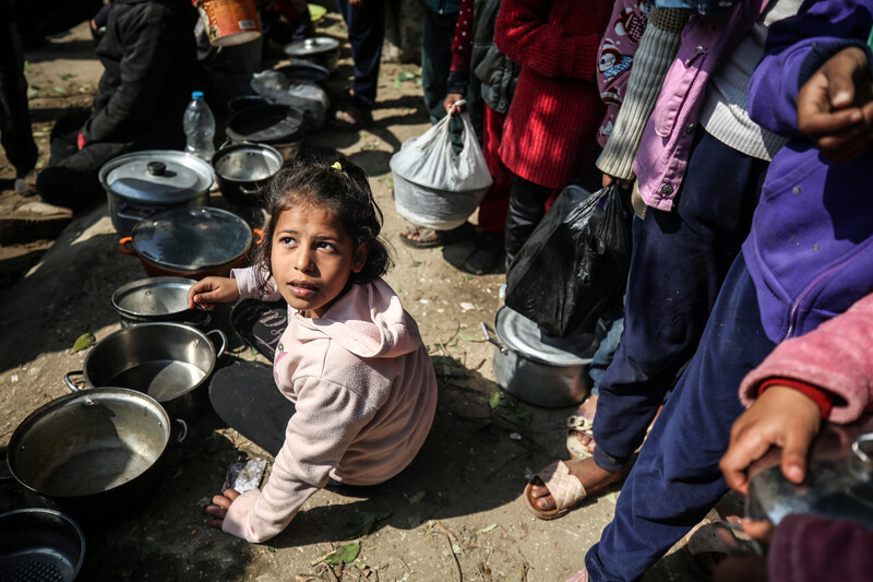 A girl sits on the ground with a tin pot in her hand. Other people are standing behind her in line