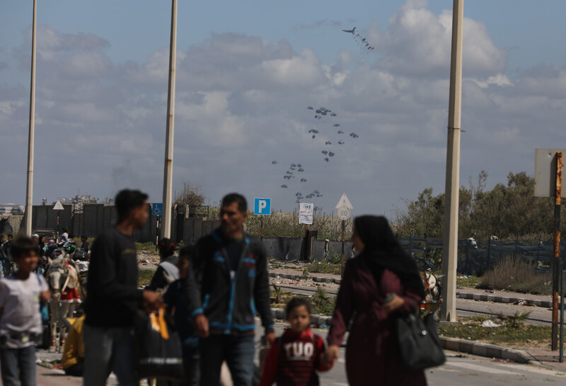 A group of people walk as parachuted packages are visible in the air behind them